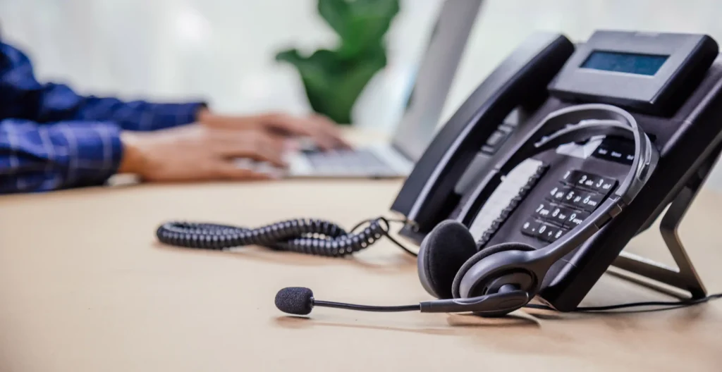 Telephone with headset sitting on a desk with someone working on a laptop in the background.
