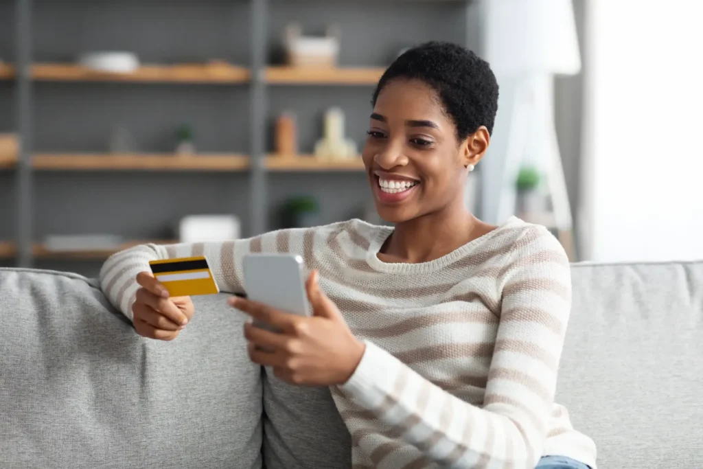 A smiling woman with holding her credit card and cell phone sitting on a living room couch.